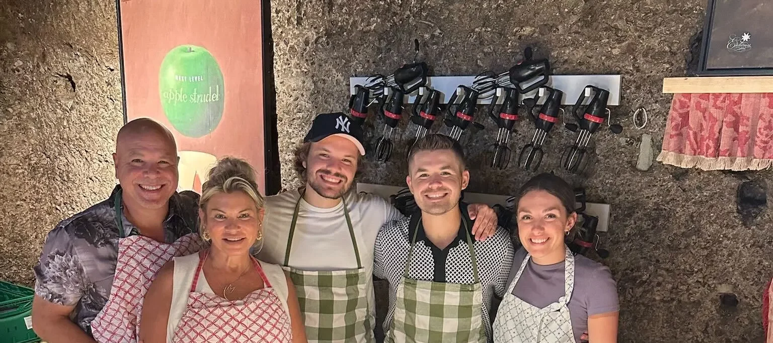 Group of five people smiling in a cozy kitchen, each wearing aprons, with a table of sliced vegetables and a rolling pin—illustrating teamwork and joy in creating a positive law firm culture.